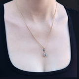 A delicate silver chain necklace with a small teardrop silver pendant worn by a model. The pendant features a small blue stone at the side and a scattering of smaller stones in the centre teardrop shape.