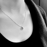 A close-up black and white photo of a  person wearing a fine chain with a round glitter pendant.