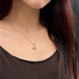 A close-up of a model wearing a delicate gold chain necklace with a small circular pendant featuring a cross/ star design, paired with a dark brown top.